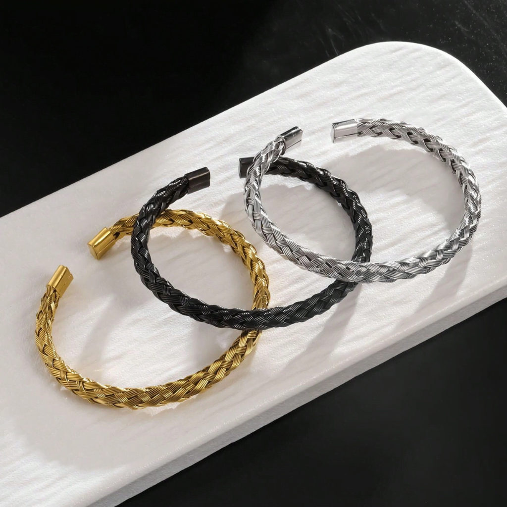 Three braided bracelets in gold, black, and silver on a white surface.